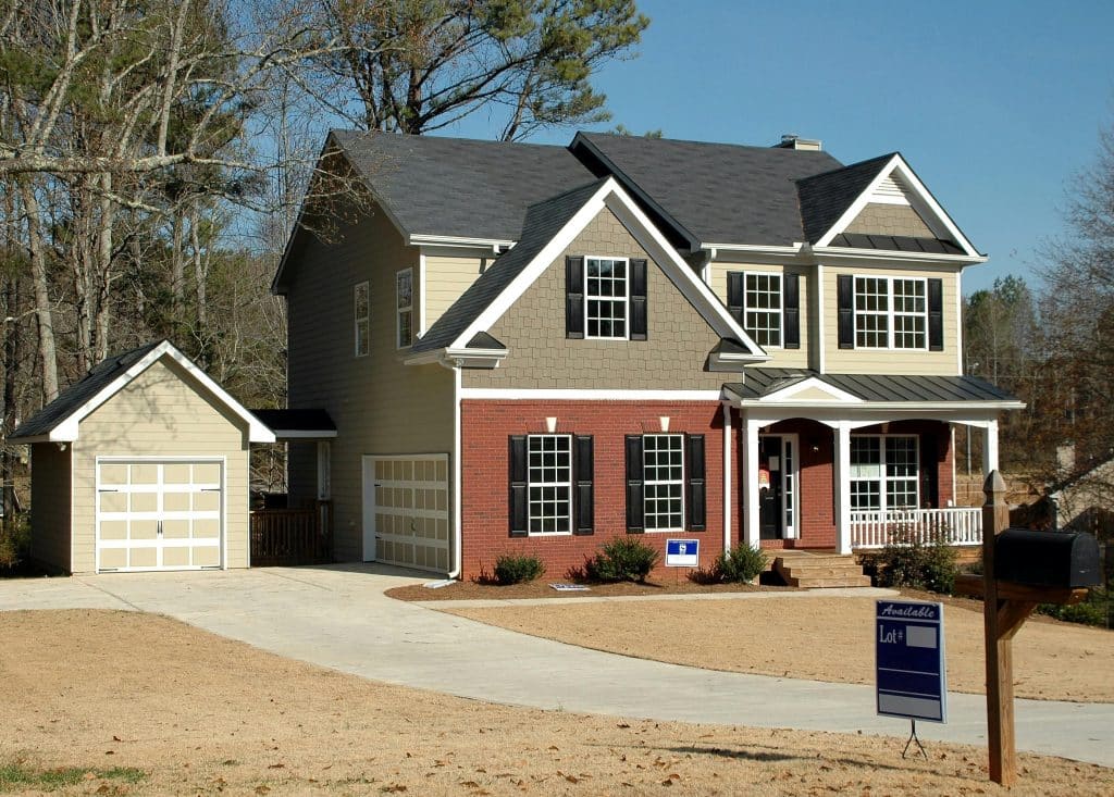Two-story suburban home with a mix of brick and beige siding, featuring black shutters, a covered front porch, and a detached garage. A "For Sale" sign is displayed in the front yard, and the driveway is clean and curved, leading up to both garages. The lawn is dry and surrounded by leafless trees, indicating a late fall or winter season.