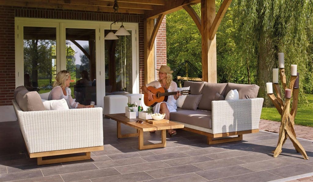 Two women relax in a cozy outdoor family living space with wicker furniture under a covered patio; one plays guitar while the other enjoys a drink.