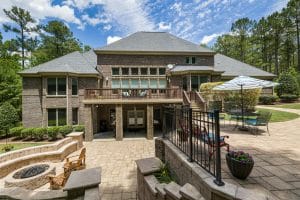 Spacious low-maintenance backyard of a large brick home featuring a multi-level patio with paver flooring, built-in fire pit, outdoor seating, dining area with umbrella, and surrounding trees for a serene, private setting.