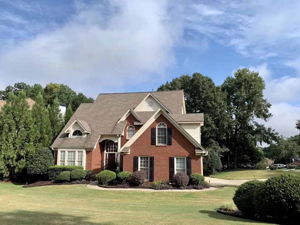 Two-story suburban brick and beige siding house with gabled roof, black shutters, and well-maintained landscaping under a partly cloudy blue sky.