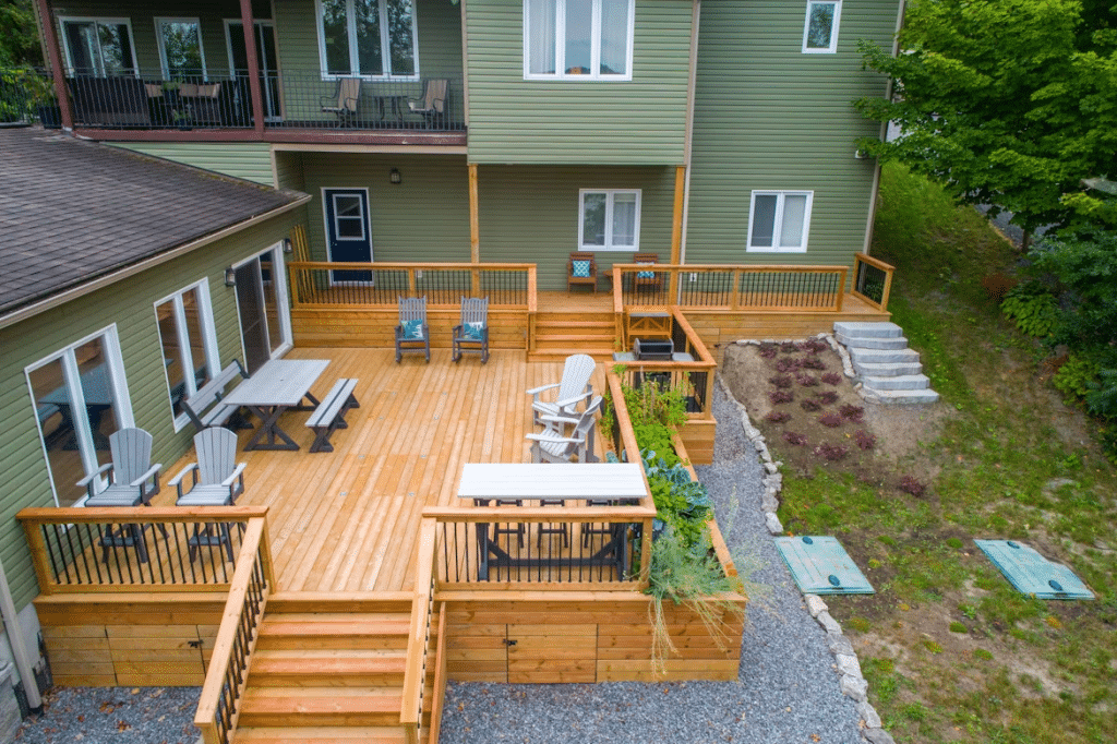 Large wooden deck with seating, planters, and stairs attached to a green house, overlooking a landscaped backyard.