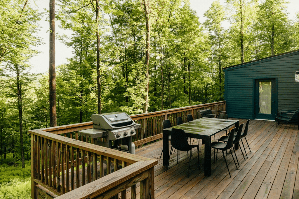 Wooden deck with outdoor dining table and grill, surrounded by dense green forest next to a modern dark house.
