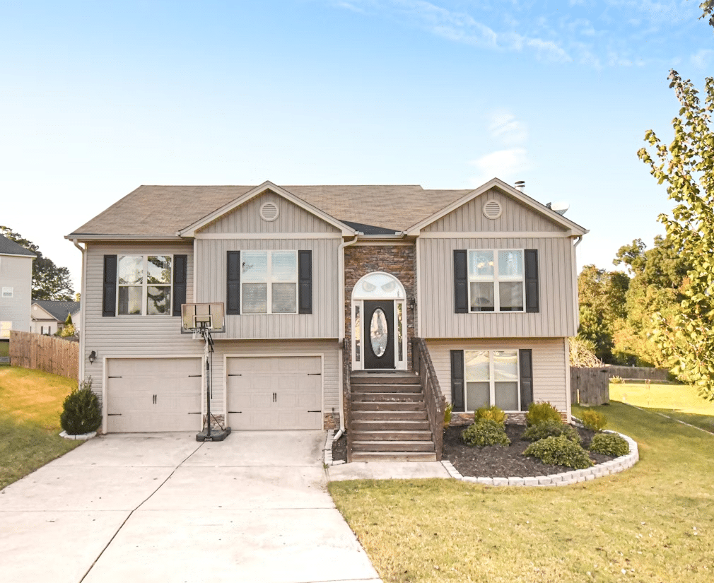 A split-level suburban home with beige siding, dark shutters, and a central stone accent surrounding a decorative front door, featuring a double garage, front steps leading to the entry, a basketball hoop, and a well-maintained lawn with simple landscaping.