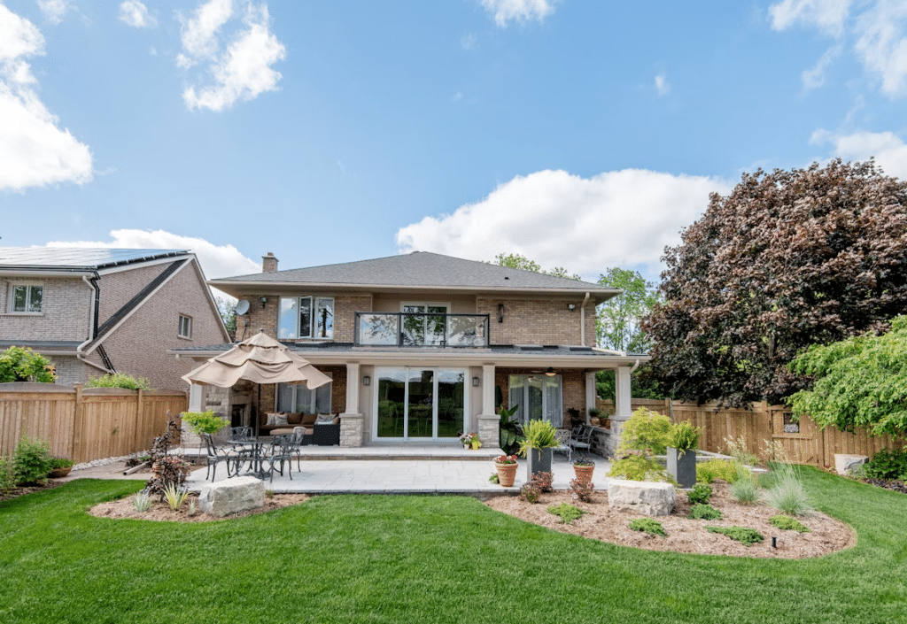 A two-story brick home with a spacious backyard, featuring a covered patio, outdoor dining area with an umbrella, landscaped garden beds, potted plants, and well-maintained green grass, all enclosed by a wooden fence and surrounded by mature trees under a partly cloudy sky.