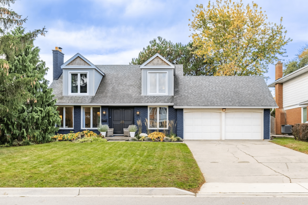 A two-story suburban home with dark blue brick siding, white trim, and a gray shingled roof, featuring a double garage, manicured lawn, front porch seating, and autumn landscaping, set against a backdrop of trees with fall foliage.