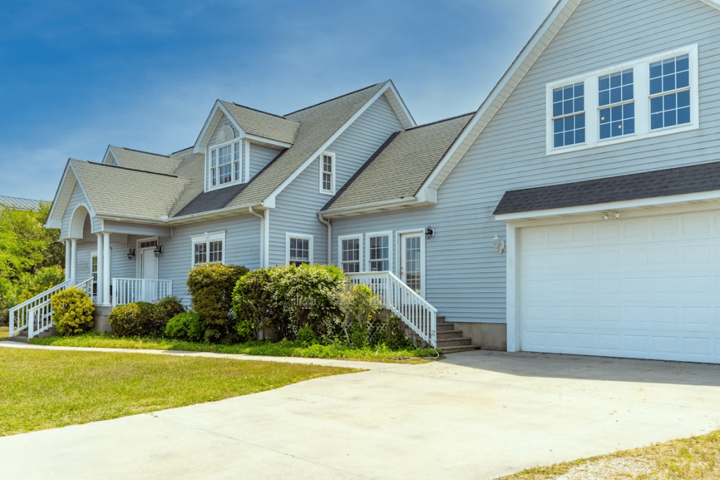 Light blue suburban house with white trim, large windows, attached garage, landscaped bushes, and a wide concrete driveway under a clear blue sky