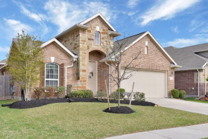 Front view of a modern brick house with stone accents, a two-car garage, landscaped yard, and young trees under a bright blue sky.