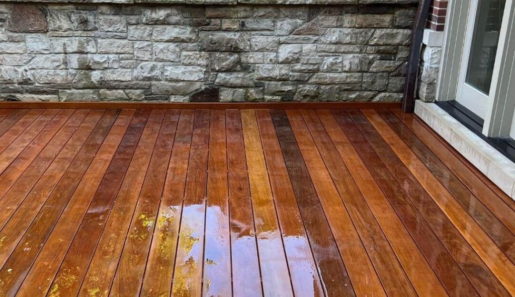 Freshly stained wood deck with a glossy finish, reflecting light and showing water resistance, set against a stone wall and adjacent to a glass door.
