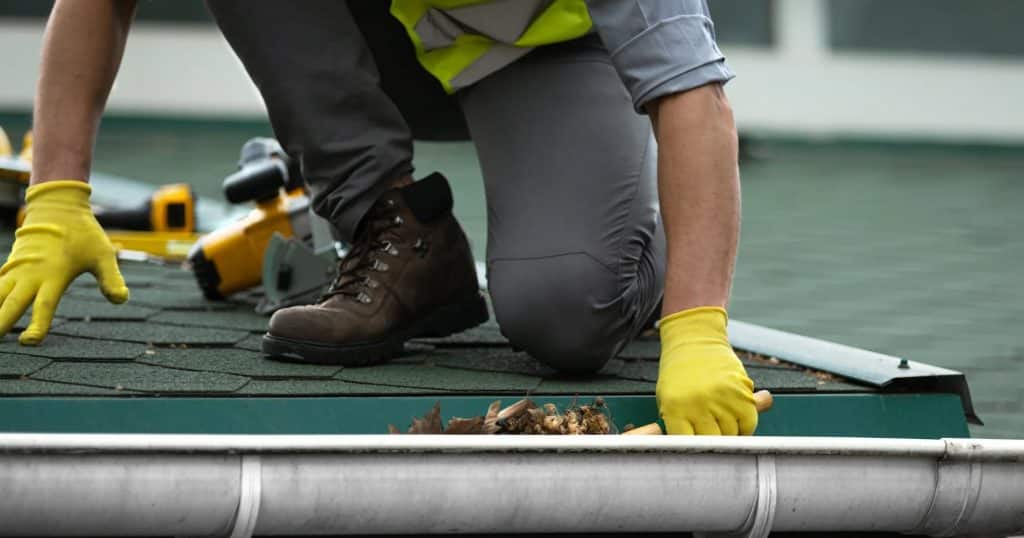 Worker wearing yellow gloves and boots kneeling on a roof while cleaning debris from a gutter.