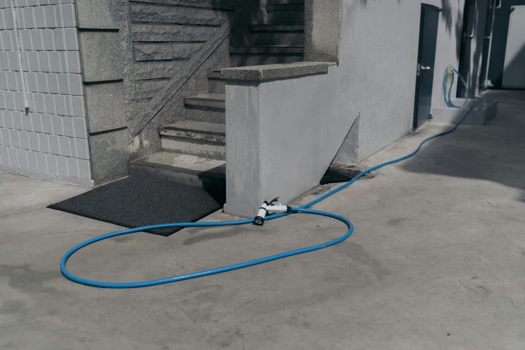 Blue garden hose with spray nozzle connected to outdoor faucet on a concrete surface near a garage.