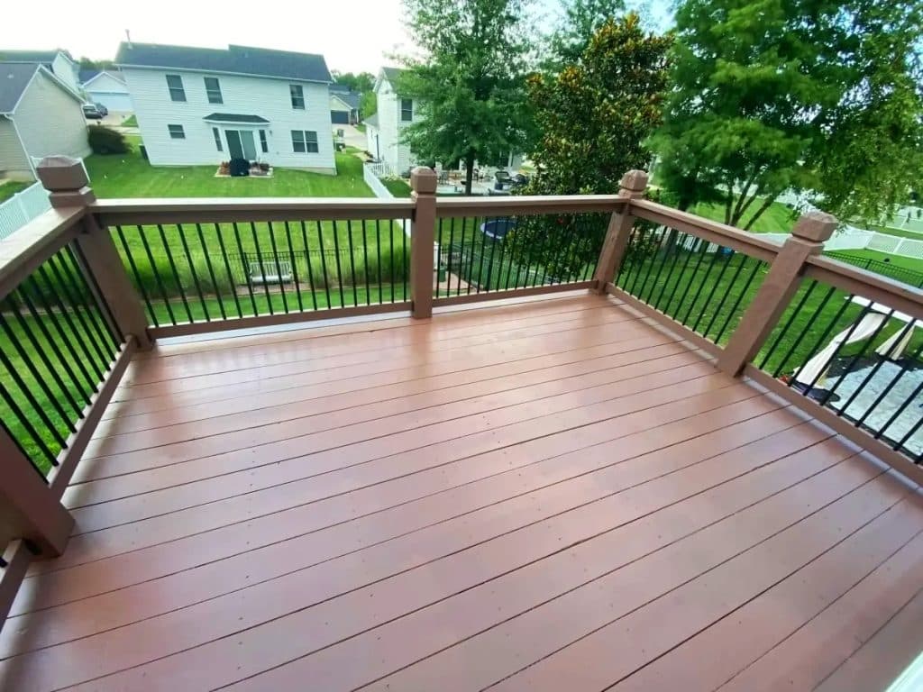 Newly stained backyard deck with brown boards and black metal railings, overlooking a green lawn and suburban neighborhood.