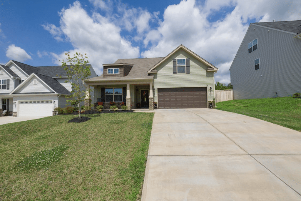 Suburban single-family home with light green siding, attached garage, covered front porch, clean gutters along the roofline, and a wide concrete driveway under a partly cloudy sky.