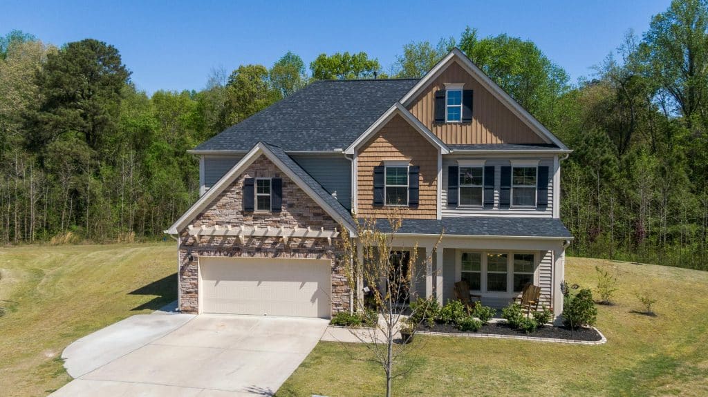 Two-story suburban home with mixed stone and siding exterior, black shutters, front porch seating, attached garage, and clean gutters along the roofline surrounded by trees.